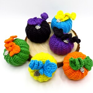 Könnte beinhalten: Sechs gehäkelte Kürbisse in verschiedenen Farben, darunter Grün, Orange, Gelb, Blau, Lila und Schwarz. Jeder Kürbis hat einen kleinen silbernen Anhänger in Form eines Baumes.