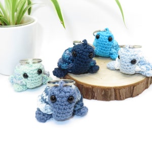 Könnte beinhalten: Fünf gehäkelte Tier-Schlüsselanhänger in verschiedenen Blautönen und Grüntönen. Die Schlüsselanhänger werden auf einer Holzoberfläche präsentiert.