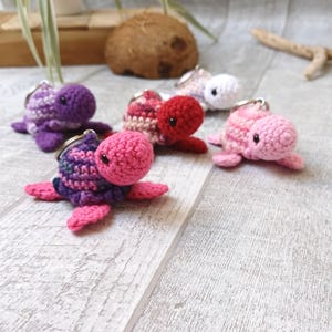 Könnte beinhalten: Fünf gehäkelte Schildkröten-Schlüsselanhänger in verschiedenen Rosatönen, Lila und Weiß. Die Schildkröten sind auf einer hellen Holzoberfläche angeordnet.