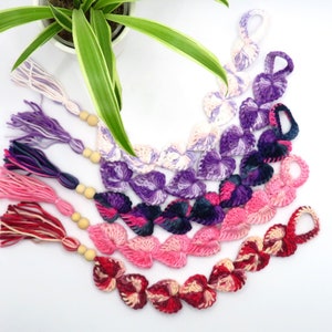 Peut inclure: Un ensemble de sept guirlandes en forme de cœur crochetées dans différentes nuances de rose, violet et bleu, chacune avec un pompon à l'extrémité.