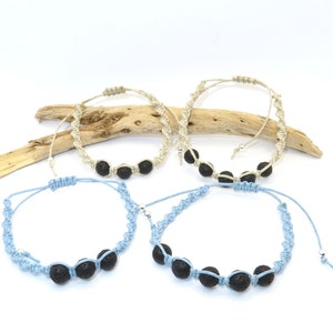 Może przedstawiać: Cztery bransoletki z makramy z czarnymi koralikami lawy. Dwie bransoletki są jasnoniebieskie, a dwie beżowe. Bransoletki mają regulowane węzły przesuwne. Na białym tle z kawałkiem drewna.