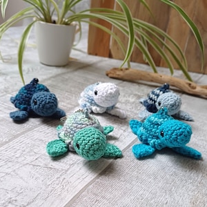 Peut inclure: Cinq porte-clés en forme de tortues de mer en crochet dans différentes nuances de bleu, vert et blanc. Les tortues sont toutes tournées vers l'avant, la tête levée et les yeux fermés.