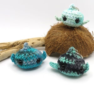 May include: Three crocheted whale keychains in different color combinations: teal and blue, green and black, and light blue and white. The keychains are sitting on a coconut shell and a piece of driftwood.