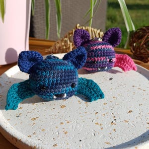 Op de afbeelding: Twee handgemaakte gehaakte vleermuisornamenten. Eén vleermuis is blauw en teal, de andere is paars en roze. Ze hebben kleine zwarte ogen en witte hoektanden. De vleermuizen liggen op een wit gespikkeld dienblad.