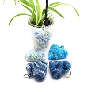 Op de afbeelding: Vier gebreide hartvormige sleutelhangers in tinten van blauw en groen. De sleutelhangers zijn in een cirkel op een wit oppervlak gerangschikt.