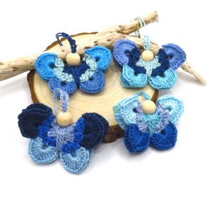May include: Four crocheted blue and white butterfly ornaments with wooden beads in the centre. The butterflies are hanging from a piece of driftwood.