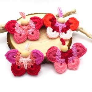 Könnte beinhalten: Vier gehäkelte Schmetterlinge mit Holzperlen in der Mitte. Die Schmetterlinge sind rosa, rot und orange.