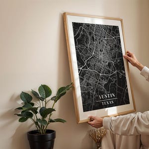 May include: A black and white map print of Austin, Texas in a wooden frame. The map is in a minimalist style with white lines on a black background. The city name and state are printed in white at the bottom of the map.