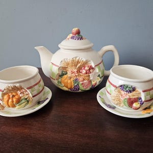 May include: A white ceramic tea set featuring a teapot and two teacups with saucers. The set is decorated with embossed images of pumpkins, wheat, and fruit. The teapot has a lid with a decorative fruit topper.