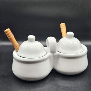 May include: A white ceramic double condiment server featuring two rounded bowls connected by a central handle. Each bowl includes a matching white domed lid and a light brown wooden spoon inserted into the side, providing a clean and functional design.