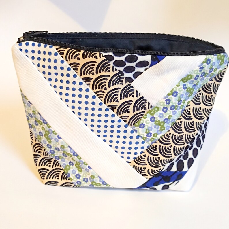 Puede incluir: Un neceser acolchado con cremallera negra e interior azul marino. La bolsa presenta un dise&ntilde;o de patchwork con tela estampada blanca, azul y beige. Los estampados incluyen lunares, flores y dise&ntilde;os ondulados.