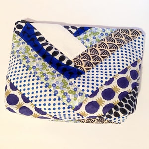 Puede incluir: Una bolsa rectangular acolchada con cierre de cremallera. La tela presenta un dise&ntilde;o de patchwork con varios patrones, incluyendo lunares azules, estampados florales y formas abstractas en azul, blanco y dorado. La bolsa mide aproximadamente 20 cm de largo.