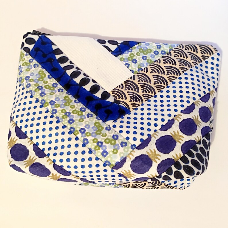 Puede incluir: Una bolsa rectangular acolchada con cierre de cremallera. La tela presenta un dise&ntilde;o de patchwork con varios patrones, incluyendo lunares azules, estampados florales y formas abstractas en azul, blanco y dorado. La bolsa mide aproximadamente 20 cm de largo.