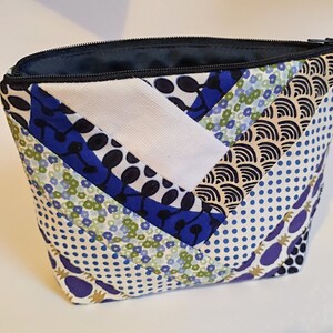 Puede incluir: Un neceser acolchado con cremallera negra e interior azul marino. El exterior presenta un dise&ntilde;o de patchwork con varios patrones, incluyendo flores azules, blanco y lunares azul marino.