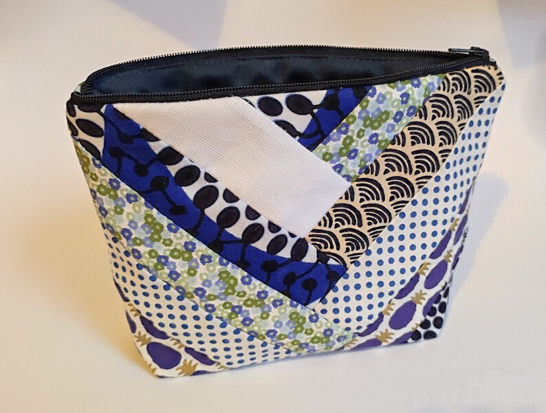 Puede incluir: Un neceser acolchado con cremallera negra e interior azul marino. El exterior presenta un dise&ntilde;o de patchwork con varios patrones, incluyendo flores azules, blanco y lunares azul marino.