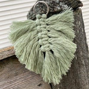 May include: A sage green macrame keychain with a silver key ring. The keychain is made of knotted string and has a fringe design.