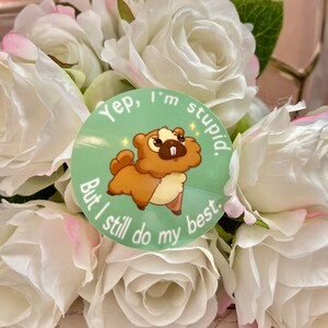 Puede incluir: Una pegatina redonda con una ilustraci&oacute;n de castor de dibujos animados en tonos marr&oacute;n y crema, sobre un fondo verde claro. El texto dice: "Yep, I'm stupid. But I still do my best." La pegatina est&aacute; rodeada de rosas blancas.
