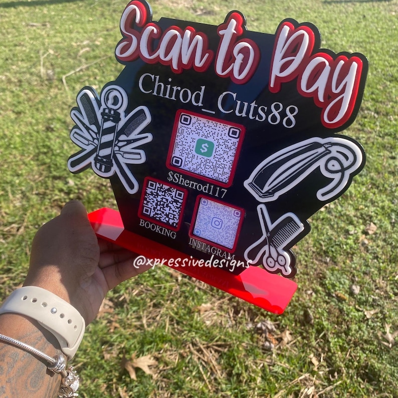 May include: A black and red sign with the text "Scan to Pay" and "Chirod_Cuts88". The sign includes QR codes for payment, booking, and Instagram. Barber shop themed with barber tools.