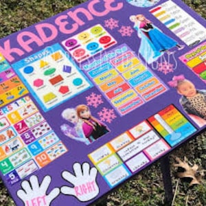 May include: A purple educational poster with the name "Kadence" at the top. The poster features colorful charts for shapes, numbers, and the alphabet, along with images of Elsa and Anna from Frozen. The poster also includes the days of the week.
