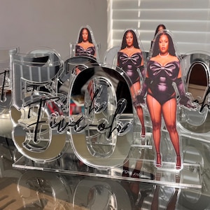 May include: A clear acrylic table centerpiece featuring the number 50 in a large font with the text Five-oh in black script layered over it. The display includes a cutout image of a person wearing a black bodysuit with a bow and gloves.