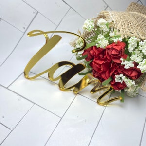 May include: Gold mirror acrylic name sign with the word "Liam" in a cursive font. The sign is placed on top of a bouquet of red and white flowers wrapped in burlap.