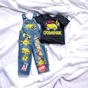 May include: Blue denim overalls with construction themed patches, including a yellow dump truck, traffic cones, and a stop sign. A black t-shirt with a yellow dump truck and the text "I'm digging Dominik" is also pictured.