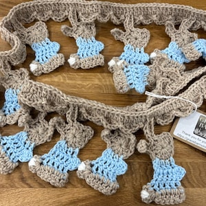 Peut inclure: Guirlande de lapins crochetés beige et bleu clair. La guirlande présente plusieurs formes de lapins avec des corps bleus et des contours beiges, reliés par une chaîne crochetée beige. Une petite étiquette est attachée.