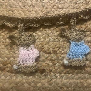 May include: Close-up of a woven basket with a decorative crochet trim. The trim features several small, handmade crochet bunny figures in shades of brown, pink, and blue. The bunnies are attached to the basket's edge, adding a whimsical touch.