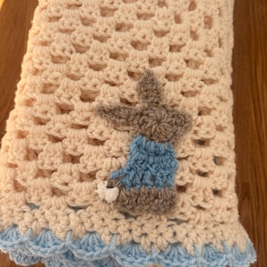 May include: A cream and light blue crochet blanket. The blanket has a repeating pattern and a bunny applique wearing a blue jacket and holding a white flower. The blanket is folded, revealing a blue scalloped edge. The blanket is made of yarn.