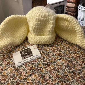 May include: A light yellow crochet hat with large, floppy ears. The hat is displayed on a floral patterned cushion. A tag with the text "GRACE BARN" is attached to the hat.