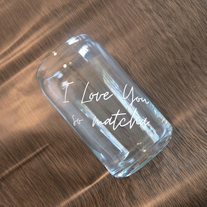 May include: Clear glass can-shaped drinking glass with the words "I Love You so matcha" in white script. The glass is empty and sits on a wooden surface. The glass has a rounded top and a rectangular base.