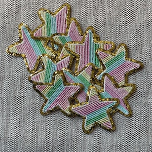 Op de afbeelding: Een groep van 10 kleurrijke stervormige patches met een gouden glitterrand. De sterren zijn gemaakt van roze, groene en blauwe gestreepte stof.