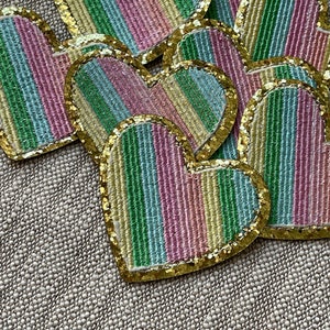 Op de afbeelding: Een stapel kleurrijke hartvormige patches met een gouden glitterrand. De harten zijn gemaakt van stof en hebben een gestreept patroon in tinten roze, groen, blauw en geel.
