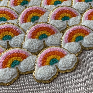 Puede incluir: Un grupo de parches de fieltro de colores arcoíris con forma de nube blanca esponjosa. Cada parche tiene un contorno de purpurina dorada y presenta un arcoíris con colores rosa, naranja, amarillo y verde.