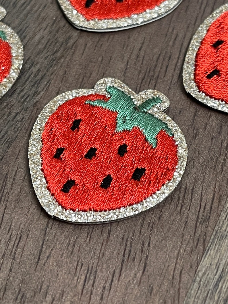 May include: A red and green embroidered strawberry patch with silver glitter around the edges.