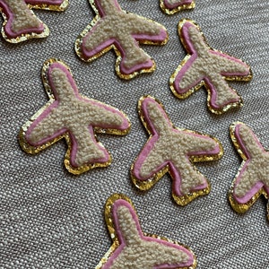 Op de afbeelding: Negen roze en gouden vliegtuigvormige patches met een pluche witte textuur. De patches zijn in een verspringend patroon op een beige stoffen achtergrond gerangschikt.