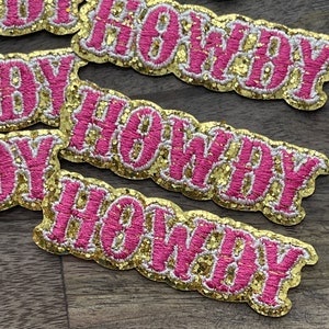 Könnte beinhalten: Nahaufnahme von vier goldenen und rosafarbenen Glitzer-Aufnähern, auf denen in einer kursivschriftlichen Schriftart "Howdy" steht.