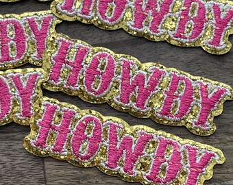 May include: Close-up of four gold and pink glitter iron-on patches that say "Howdy" in a cursive font.