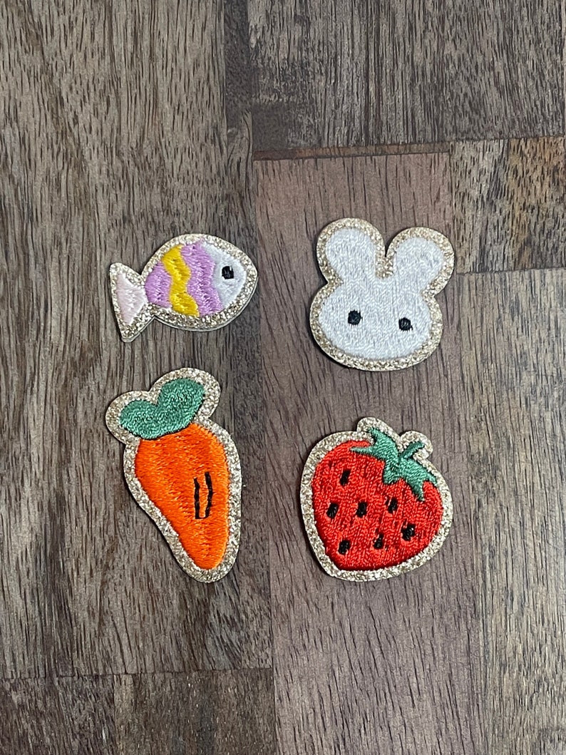 May include: Four embroidered iron-on patches with a gold glitter outline. The patches feature a pink and yellow fish, a white bunny, an orange carrot, and a red strawberry.