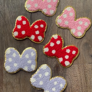 Puede incluir: Seis lazos de tela de fieltro con ribete dorado y lunares blancos. Los lazos son de tres colores: rosa, rojo y morado.