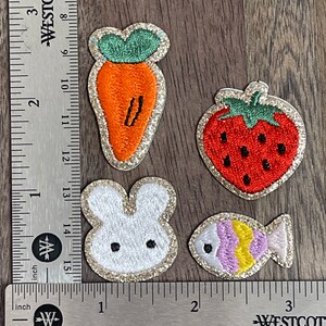 May include: Four embroidered iron-on patches with a glitter finish. The patches feature a carrot, a strawberry, a bunny rabbit, and a fish.
