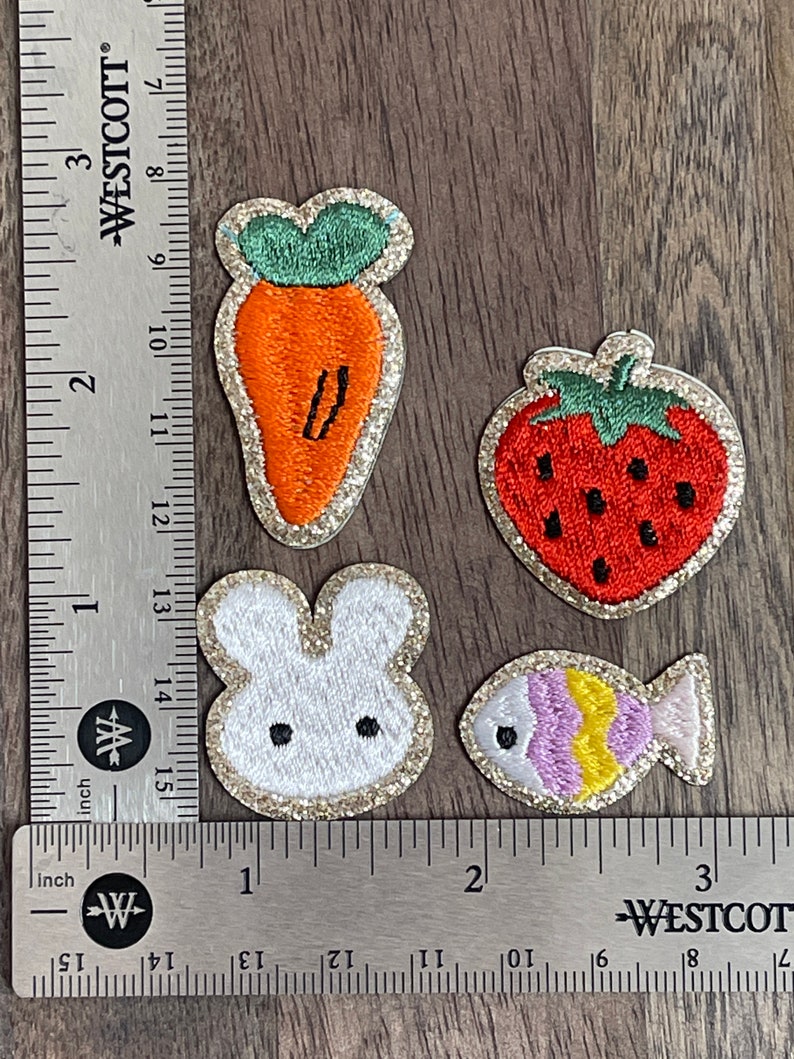 May include: Four embroidered iron-on patches with a glitter finish. The patches feature a carrot, a strawberry, a bunny rabbit, and a fish.