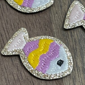 May include: Two purple, yellow, and white glittery fish with black eyes. The fish are embroidered with a wavy pattern.