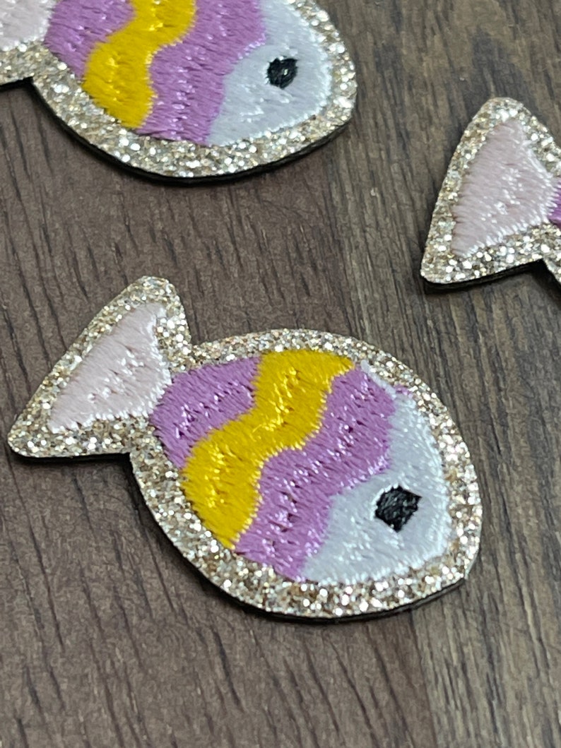 May include: Two purple, yellow, and white glittery fish with black eyes. The fish are embroidered with a wavy pattern.