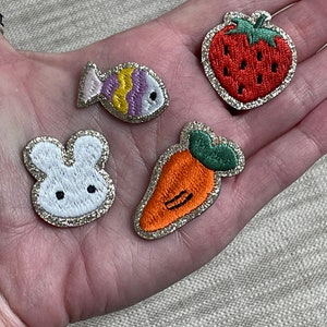 May include: Four embroidered iron-on patches with a glitter background. The patches feature a white bunny, a red strawberry, an orange carrot, and a purple and yellow fish.