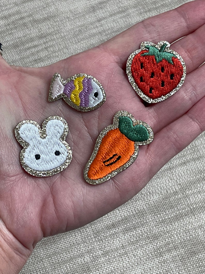 May include: Four embroidered iron-on patches with a glitter background. The patches feature a white bunny, a red strawberry, an orange carrot, and a purple and yellow fish.