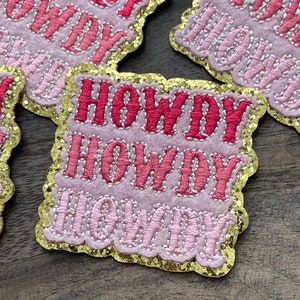 Puede incluir: Tres parches de fieltro rosa y rojo con ribete de purpurina dorada. Cada parche dice "Howdy" en una fuente en capas.
