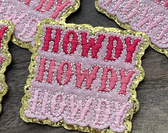 May include: Three pink and red felt patches with gold glitter trim. Each patch says "Howdy" in a layered font.