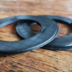 May include: Two black plastic rings, probably for a 3D printer, are stacked on top of each other on a wooden surface.