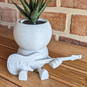 Puede incluir: Maceta de cerámica blanca con forma de persona tocando una guitarra, con una planta suculenta verde en su interior. La maceta tiene una superficie texturizada y el texto "ohne Pflanze" es visible en la esquina superior derecha de la imagen.